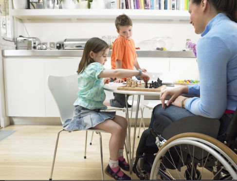 A woman using a wheelchair is at a table with two young children at home playing a game.