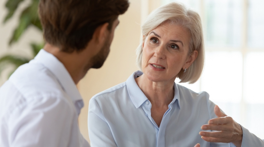 Older professional woman with fair hair talking to a young man.