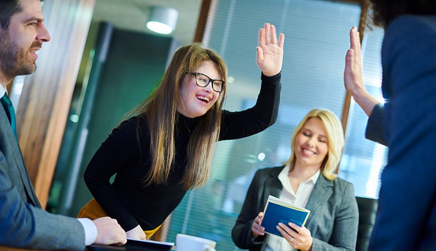 Two colleagues are raising hands for a high five. One is partially out of frame and the other is a young woman with long blonde hair and Down Syndrome.  A male colleague is smiling and  a woman is also smiling and looking down at a book.