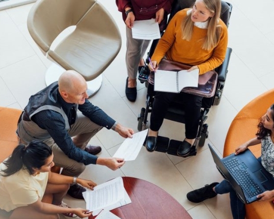 Four people sitting down, including one wheelchair user, and one person standing. They are engaged in discussion.