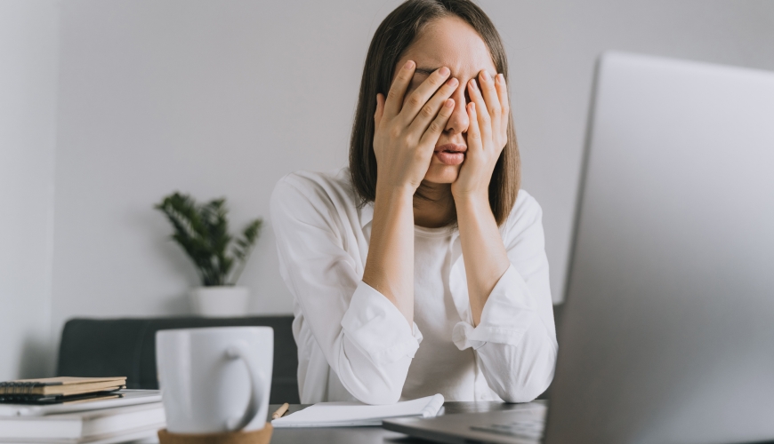 Female corporate woman sitting at desk hiding her face with hands.