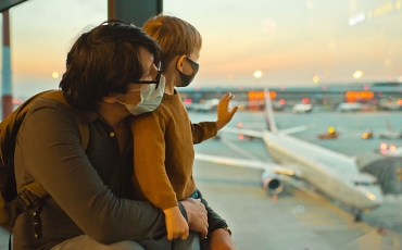 Father and small child at airport, looking out the window at waiting planes. Both are wearing covid masks. 