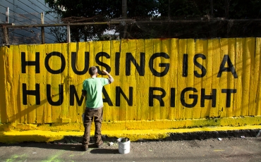 man paints 'housing is a human right' on yellow wooden fence