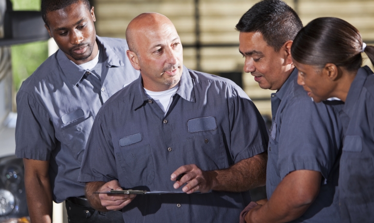 "Team of multi-ethnic mechanics. Focus on Caucasian man holding clipboard.""