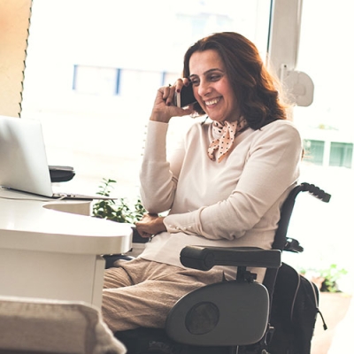 A woman in a wheelchair is smiling while she is on a phone call. She is working from home on her laptop.