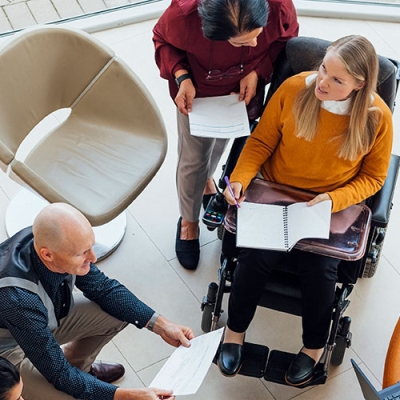 A bird’s eye view of a group of people sitting in a circle with laptops and notebooks. Some people are on couches, and others are on chairs. One woman is in a wheelchair.