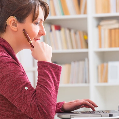 A woman sitting at a desk who is working on her computer. She is dressed in red and has a hearing device.