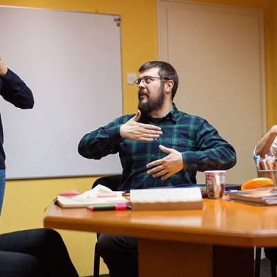 A group of men and women are in a staff meeting. They are all communicating  together through sign language.