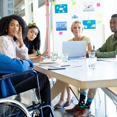 A group of colleagues sitting around a table from various ethnicities and ages.  One man is sitting in a wheelchair.