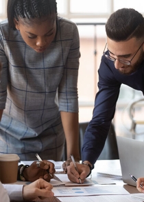 A group of employees lean around a table, collaborating on a document together.