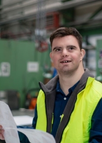 A man with Down Syndrome in an industrial working environment holding some paperwork and wearing a safety vest.